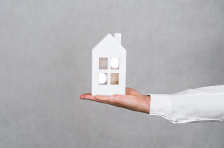 A hand in a white shirt holds a small white house model against a neutral gray background, symbolizing real estate, home ownership, and property investments.の写真素材