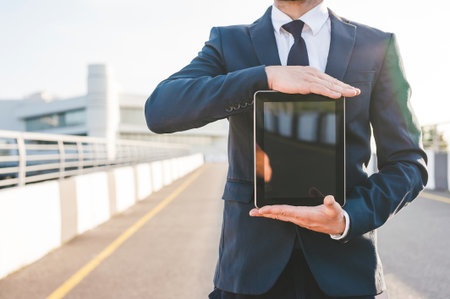 Man in formal suit holding a tablet with a blank screen outdoors. Ideal for showcasing apps, web design, or digital services in a business context.の写真素材