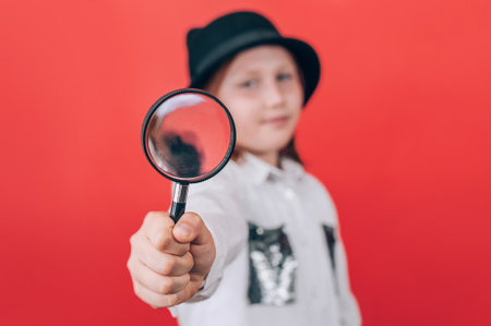 A young child, wearing a hat, holds a magnifying glass towards the viewer, symbolizing curiosity, investigation, and exploring potential future professions or interests.の写真素材