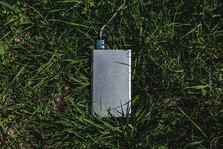 Silver power bank with cable lying on green grass. Outdoor charging setup blends modern technology...
