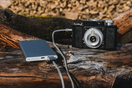 Close-up of a vintage camera connected to a modern power bank via cable, charging outdoors on wooden logs. Perfect concept for technology, photography, and charging portable solutions.の写真素材