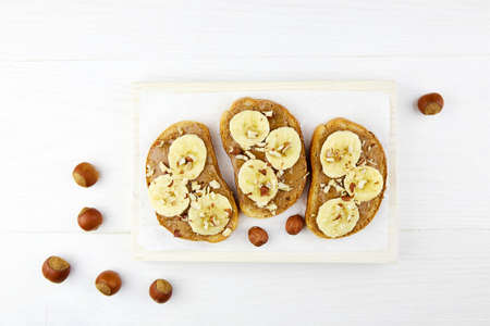 Toasts with hazelnut paste, banana and hazelnuts on white wooden background. Diet natural breakfast.