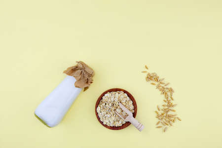 Oat milk in a bottle, oat flakes in a ceramic bowl on a yellow background. Vegetable milk.