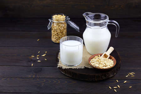 Oatmeal milk in a jug and a glass, oats in a jar, oatmeal flakes in a bowl on a dark wooden background. Vegetable milk. Vegan lactose free.の写真素材