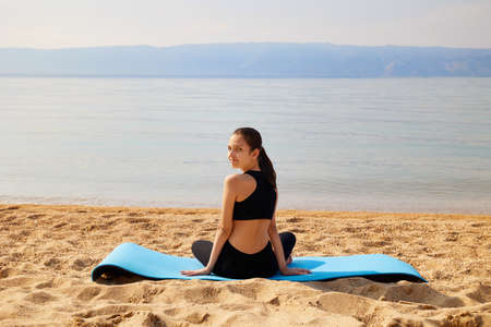 A sporty girl is sitting on a yoga mat on a sandy beach on the shore of a lake. Yoga time.
