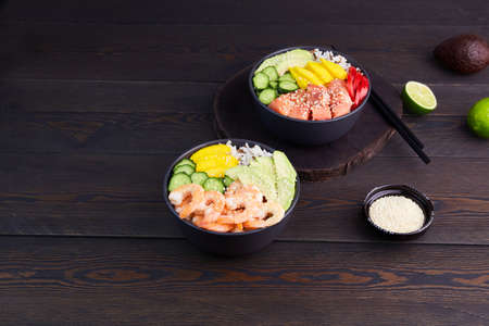 Poke bowl with salmon, shrimps, avocado, rice, cucumber, mango, pepper and sesame seeds, soy sauce on dark wooden background.の写真素材