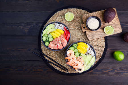 Poke bowl with salmon, shrimps, avocado, rice, cucumber, mango, pepper and sesame seeds on dark wooden background. Top view.の写真素材