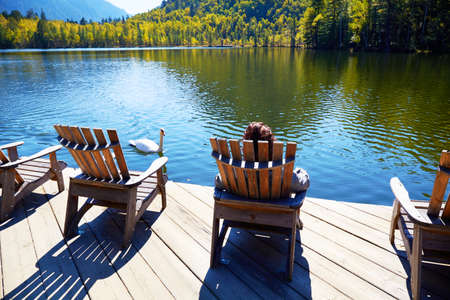 A girl is resting on a wooden sun lounger on the shore of an Emerald Lake on a warm autumn day. A white swan is swimming nearby. Travel and outdoor recreation concepts.の写真素材