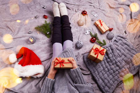 A young girl in a knitted gray wool sweater holds a gift box. Sitting on the bed, next to gifts, Christmas toys. Christmas lights bokeh. Cozy atmosphere of Christmas, holiday.の写真素材