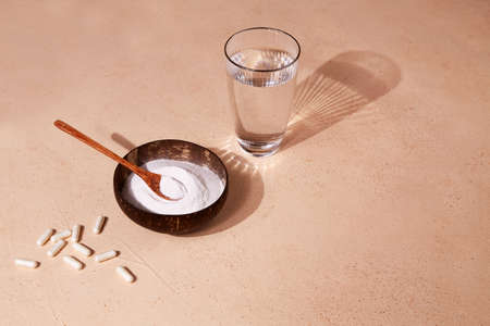 Collagen protein powder in a bowl, capsules and glass of water on a beige background. A food...