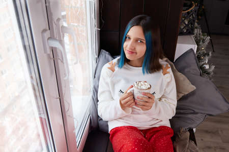 A pretty girl in pajamas is sitting on the windowsill with a cup of cocoa with marshmallows. The room is decorated with a Christmas tree, toys. New year.の写真素材