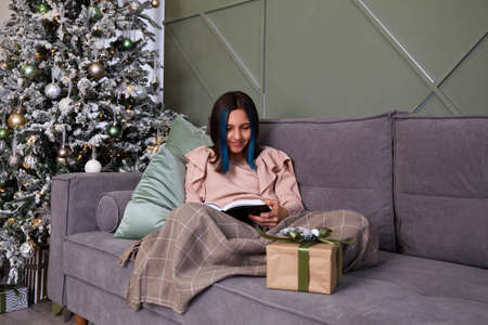 A cute girl is reading a book on the sofa, covered with a blanket. Christmas tree with toys and gifts.の写真素材