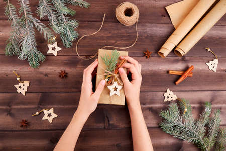 Women's hands are packing a Christmas gift. Fir branches, gift box, wrapping paper, cinnamon sticks, wooden Christmas tree toys.の写真素材