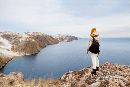 A young girl in warm clothes with a hiking backpack is standing at the cliff, admiring nature....