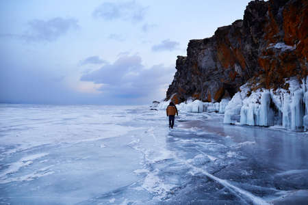 Frozen Lake Baikal at sunset. A male traveler walks on the transparent ice of the lake. The rocks are covered with snow and icicles. Winter hiking, outdoor activities.の写真素材