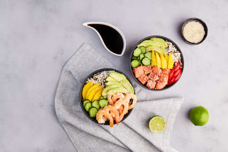 Poke bowl with shrimps, tuna, avocado, rice, cucumber, mango, pepper and sesame seeds on gray background. Hawaiian ahi, pokebowl, a diet meal with seafood. top view.の写真素材