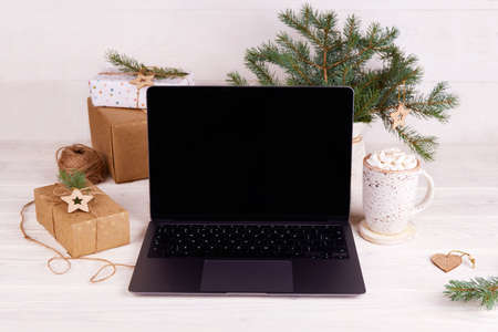 Laptop screen, cocoa mug with marshmallows, gift box, Christmas tree branches on a white wooden background. Shopping online for the Christmas holidays. Mock upの写真素材
