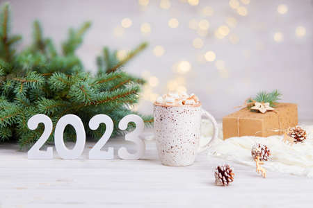 Happy New Year greeting card 2023. A mug of cocoa with marshmallows, a gift in kraft paper, white wooden numbers 2023 on the background of a beautiful bokeh. christmas holiday background.の写真素材