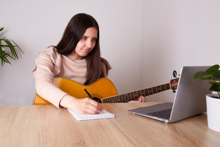 A young girl is learning to play the guitar. A teenager is sitting at a table in front of a laptop, watching an online lesson and writing it down in a notebook.の写真素材