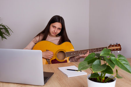 A young girl is learning to play the guitar. A teenager is sitting at a desk in front of a laptop and listening to an online lesson. distance education.の写真素材