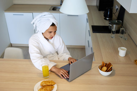 A girl in a bathrobe is working at a laptop in the kitchen.の写真素材