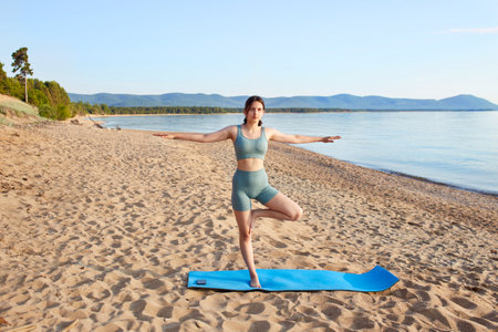 Sporty girl does yoga on sandy beach by the sea. The athlete stands on one leg, with her arms spread to the sides. Balance and pose trainingの写真素材