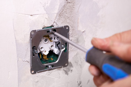 A worker installs an electrical outlet into a wall using a screwdriver. The apartment is undergoing...