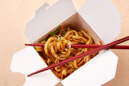 Delicious noodles with colorful vegetables are neatly packed in a white takeout box. Red chopsticks are poised above the dish, ready for enjoyment. Top view.の写真素材