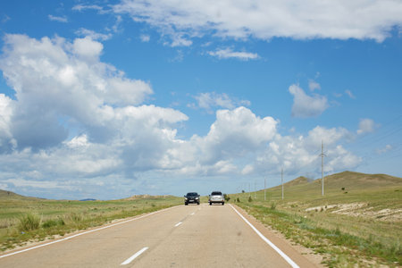 Two cars navigate a winding road surrounded by vast grasslands and rolling hills. The sky is clear with large, fluffy clouds adding to the scenic view. Summer tripの写真素材