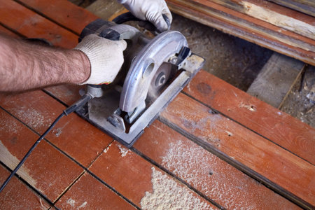 Craftsman operates a circular saw to cut through wooden planks in a well-lit workshop. Dust rises as precision work is conducted, showcasing skill and focus in carpentry.の写真素材