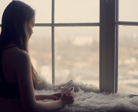 Woman in the morning. woman with neat body is holding a cup with hot tea or coffee and looking at the sunrise standing near the window in her home and having a perfect cozy morningの写真素材