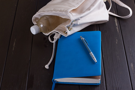 White leather backpack, blue notebook and pen on a black wooden backgroundの写真素材
