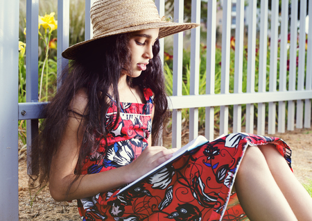 Adorable girl in dress and hat reading book in summer park.の写真素材