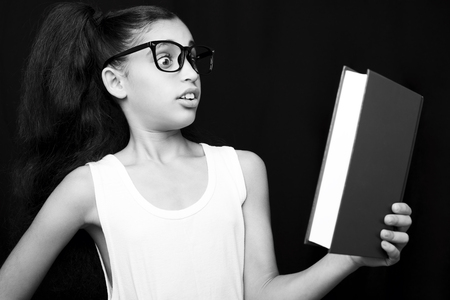Adorable girl studying with eyeglasses and book in hand on black backgroundの写真素材
