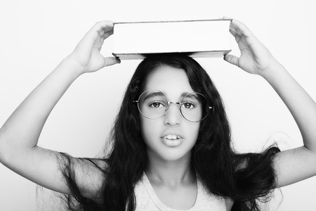 Adorable girl studying with eyeglasses and book on the head on white backgroundの写真素材