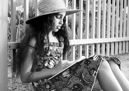 Adorable girl in dress and hat reading book in summer park.の写真素材