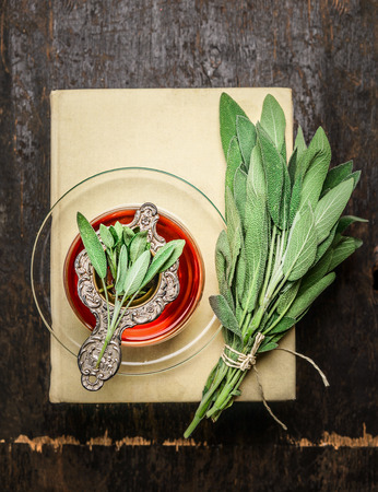 Cup of herbal sage tea with old strainer, books and bunch of herbs on rustic wooden background, top viewの写真素材