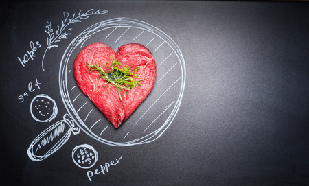 Heart shaped chop of meat on black chalkboard with  painted pan and ingredients, top view, place for text. For Meat lovers and eaterの写真素材