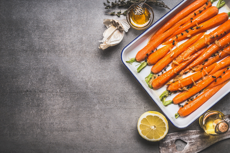 Roasted Baby Topped carrots on baking sheet with thyme, honey, garlic and lemon on gray concrete background, top view, border. Healthy root vegetables conceptの写真素材
