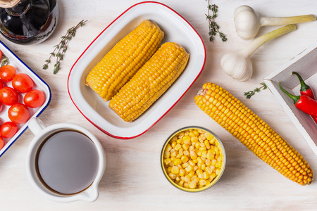 Ingredients for corn soup or stew: ear of corn, canned and cooked corn , seasoning and vegetables broth on white wooden background, top viewの写真素材