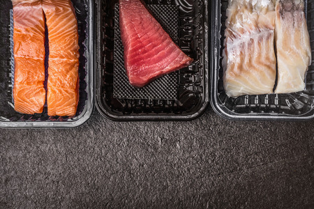 Selection of raw colorful fish fillets : salmon, tuna and codfish in plastic boxes on dark rustic background, top view, border, place for text. Healthy Seafood conceptの写真素材