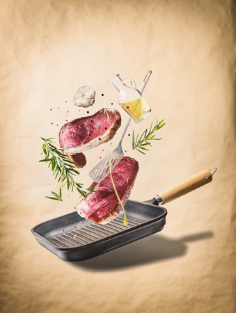 Flying raw beef meat steaks, with herbs, oil and spices with grill pan and kitchen utensils, at natural beige background, front view. Flying  food conceptの写真素材