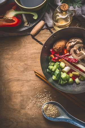 Wok pot with vegetarian asian cuisine ingredients for stir fry with chopped vegetables, spices, sesame seeds and chopsticks on rustic wooden background, top view. Chinese or Thai food cooking conceptの写真素材