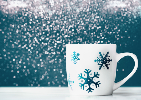 White mug with snowflakes on table at blue background with bokeh, front view concept. Winter holidays layoutの写真素材