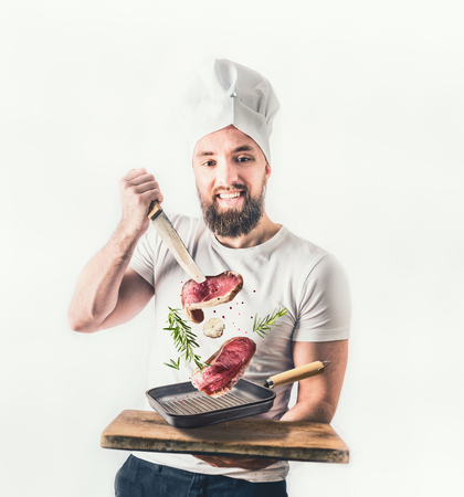 Young cheerful cook man holding a pan of steak and knife flying in the air on light background. Meat food and cooking conceptの写真素材
