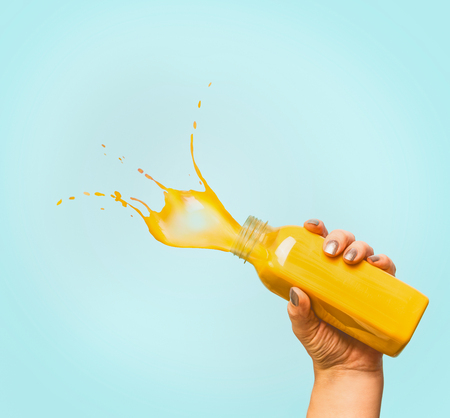 Female hand holding bottle with yellow splash summer beverage: smoothie or juice at blue background, copy spaceの写真素材