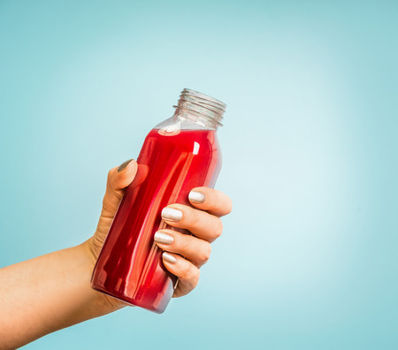 Female hand holding bottle with red summer drink: smoothie or juice at blue background.