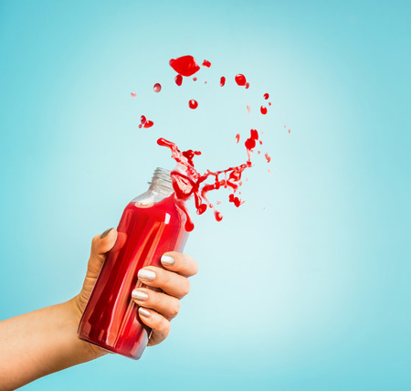 Female hand holding bottle with red splash summer beverage: smoothie or juice at blue background.の写真素材