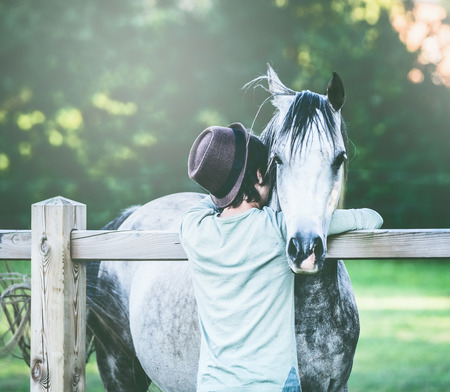 Young guy with hat hugs his gray horseの写真素材