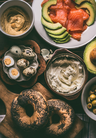 Breakfast preparation with bagel bread, salmon, avocado , fresh cheese, hummus and cooked quail eggs on dark rustic wooden background, top view.の写真素材